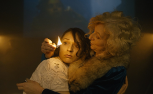 Starsza kobieta trzymając w dłoni zapalona zapałkę przytula młodszą, która wpatrzona jest w płomień.